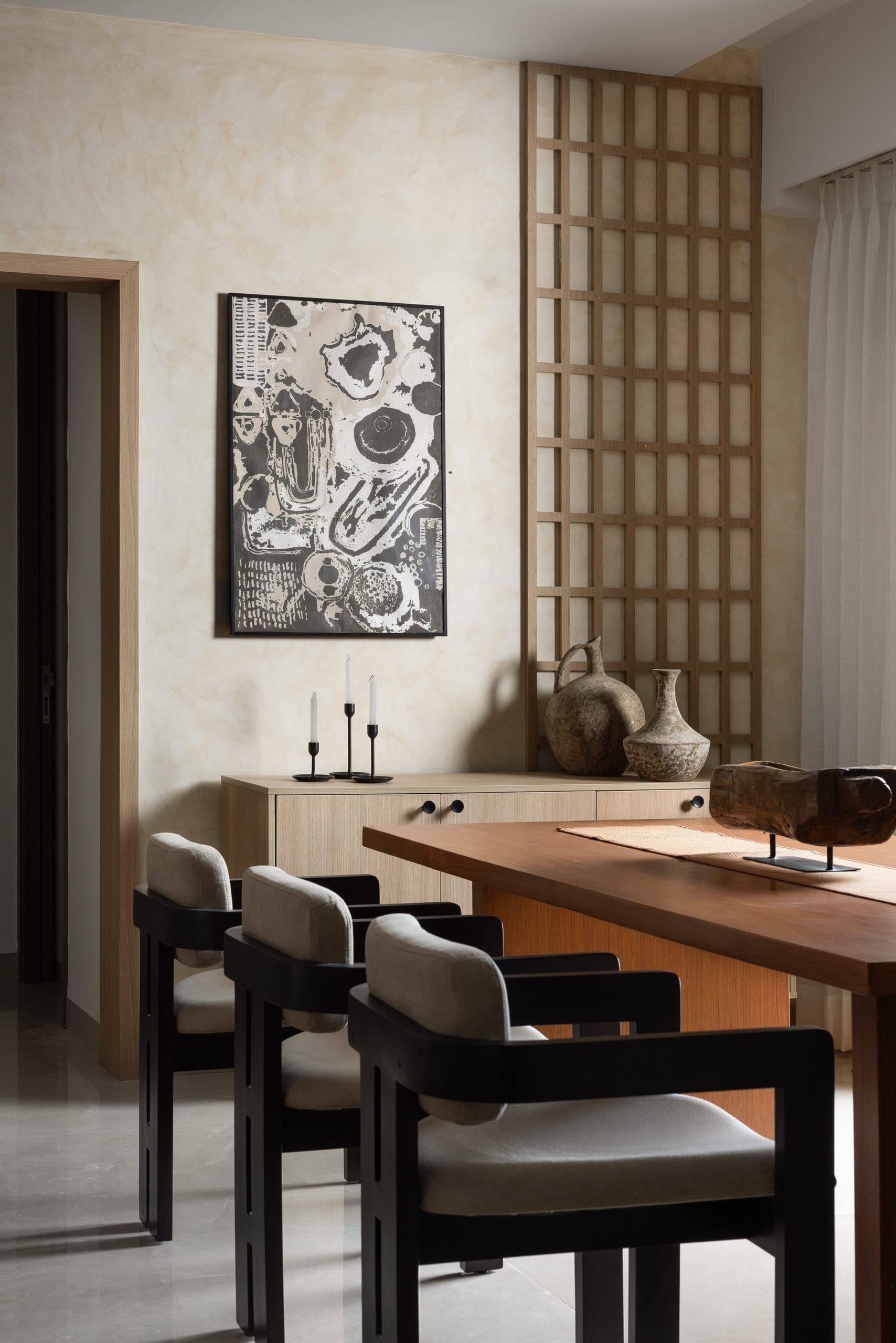 The dining area continues the home's earthy material language. A solid wood table is paired with black-framed armchairs upholstered in natural linen, their squared geometry echoing the graphic lattice screen that divides the dining and passage zones. Antique-finish ceramic vessels and taper candles complete the tableau.