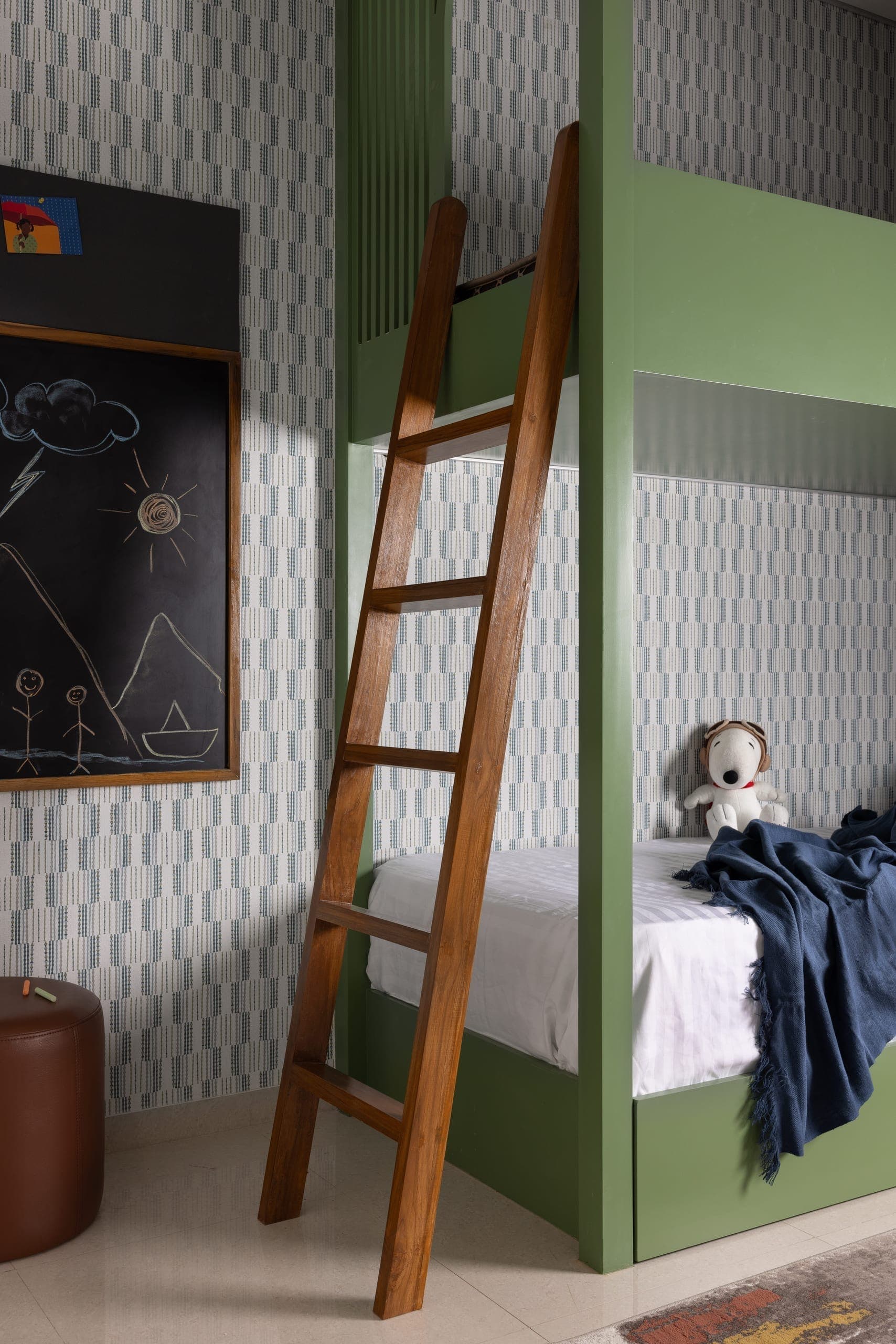 A custom olive green bunk anchors the room, with a teak ladder and patterned wallpaper adding a light, playful layer. A framed chalkboard panel allows the space to evolve over time.