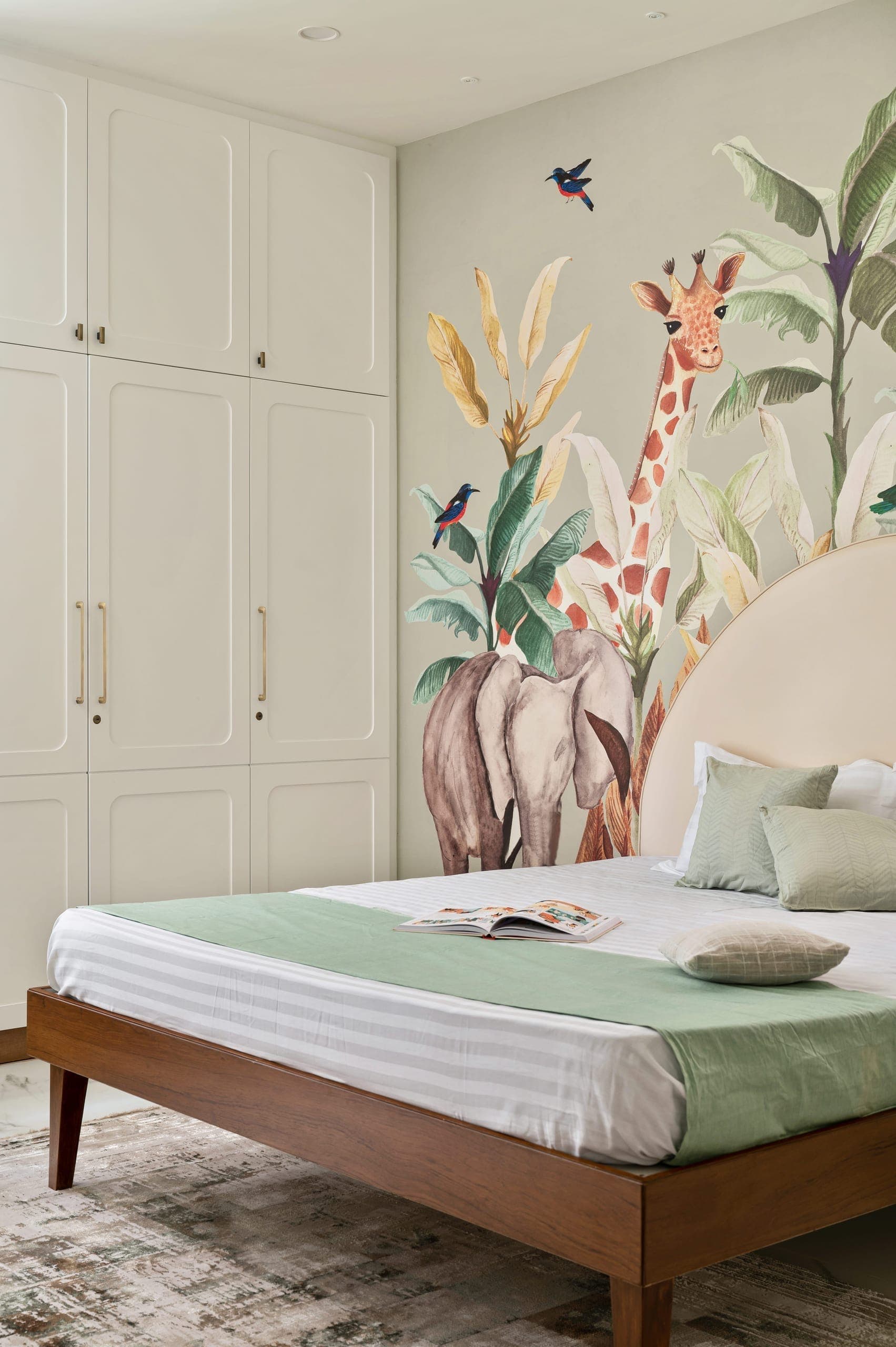 The bedroom is defined by a neutral palette of white and warm wood, creating a calm and inviting base. Soft creams and subtle greens add lightness without overpowering the space. The headboard wall introduces a playful note through a patterned wallpaper, bringing character while maintaining the room's overall restraint.
